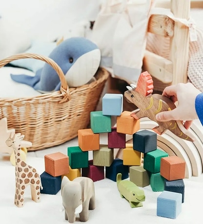 Main jouant avec un dinosaure en bois, près de blocs colorés, d'une girafe et d'un éléphant en jouets écologiques.