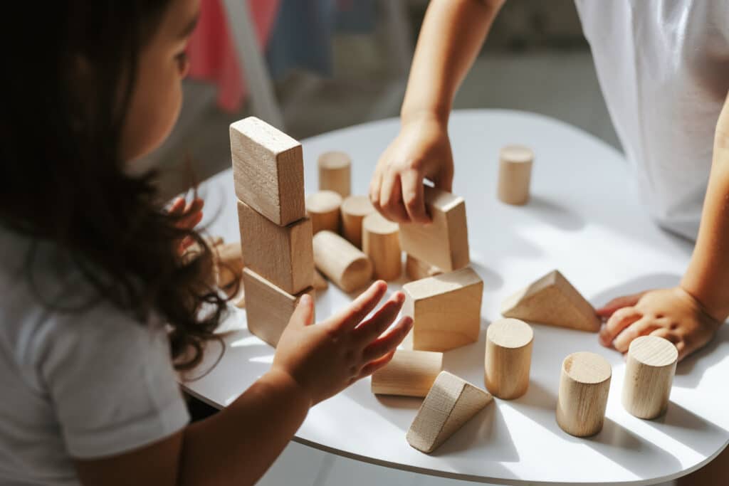 Deux enfants jouent ensemble avec des blocs de construction en bois clair empilés sur une table blanche, leurs mains sont visibles.