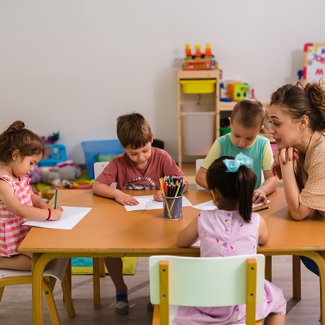 Enseignante et quatre jeunes enfants dessinant à une table en bois dans une salle de classe remplie de jouets.