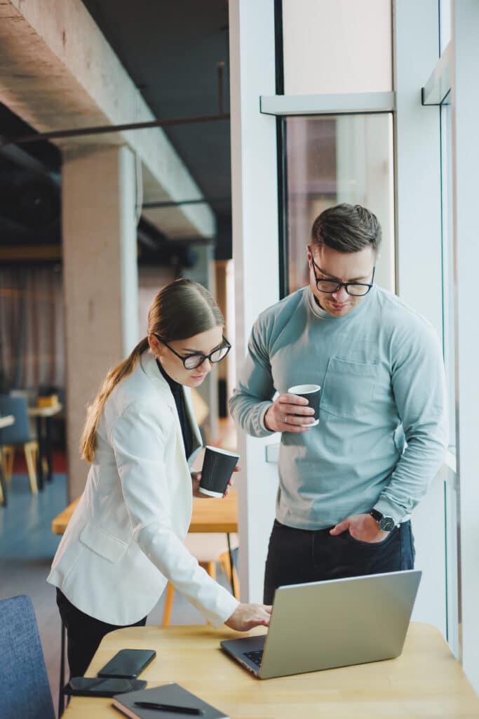 Deux professionnels, un homme et une femme portant des lunettes, consultent un ordinateur portable sur un bureau, chacun tenant une tasse de café.