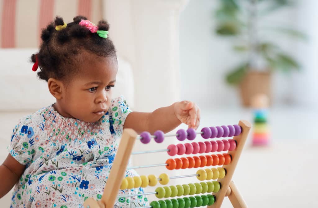Jeune enfant métisse concentrée jouant avec un boulier en bois coloré à perles violettes, roses, oranges, jaunes et vertes.