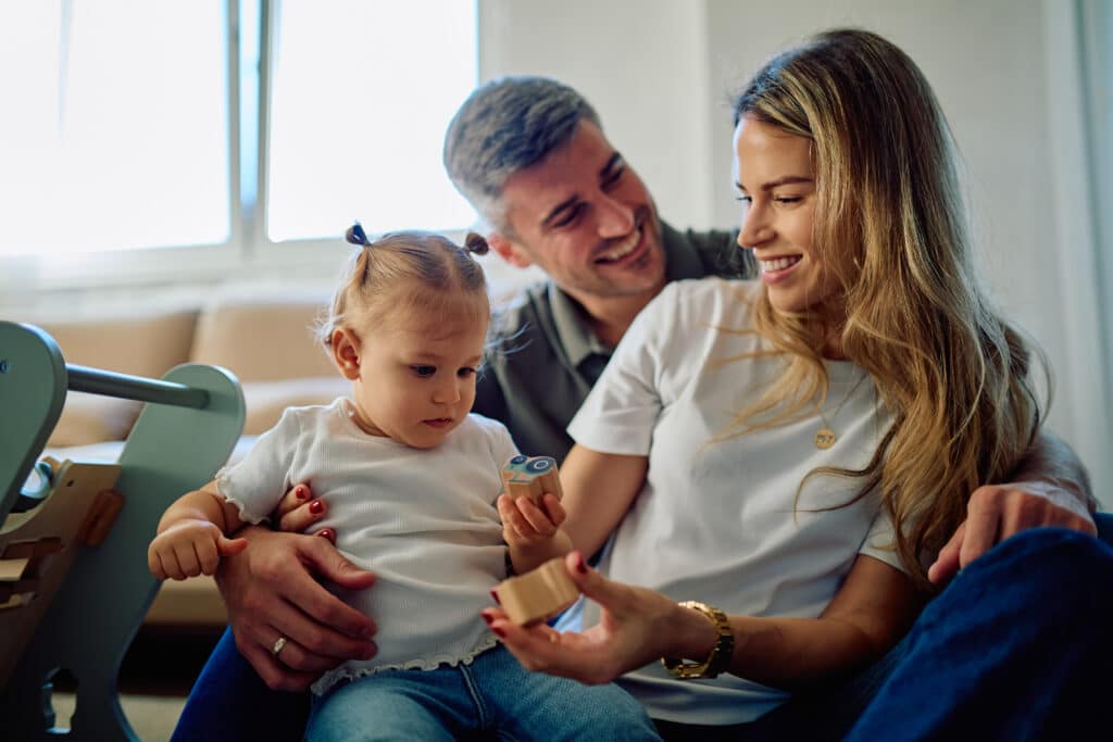Un père et une mère souriants jouent avec leur jeune fille blonde qui tient des blocs de bois dans un intérieur lumineux.