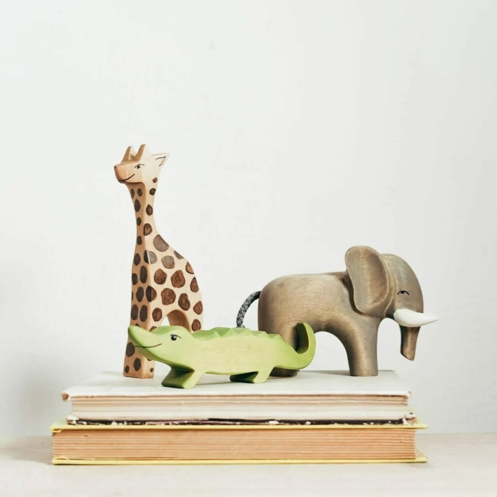 Figurines en bois d'animaux de la savane : girafe tachetée, crocodile vert et éléphant gris, posées sur une pile de livres clairs.