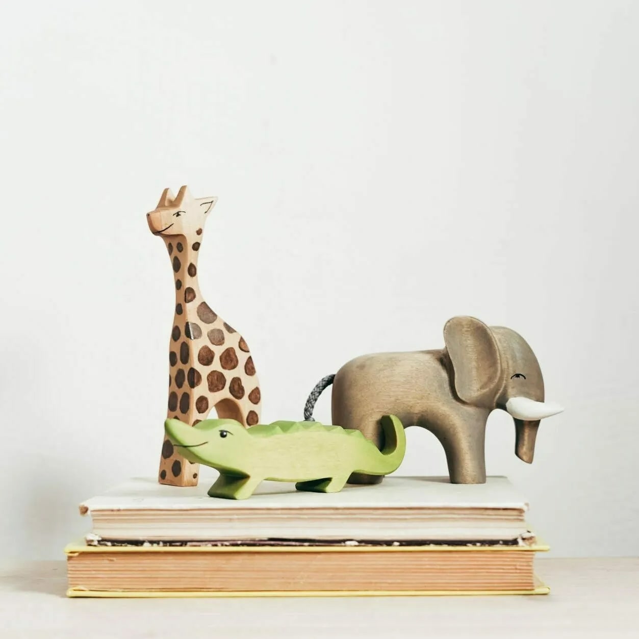 Figurines en bois d'animaux de la savane : girafe tachetée, crocodile vert et éléphant gris, posées sur une pile de livres clairs.
