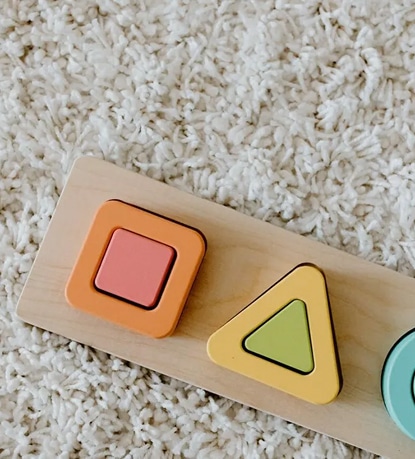 Puzzle en bois pour bébé posé sur un tapis à poils longs blanc, montrant des pièces carrée orange et triangulaire jaune.