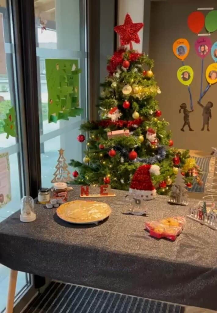Table décorée pour Noël avec sapin, tarte, pot de Nutella, et décorations rouges et blanches. Des lettres formant "N E" sont visibles.